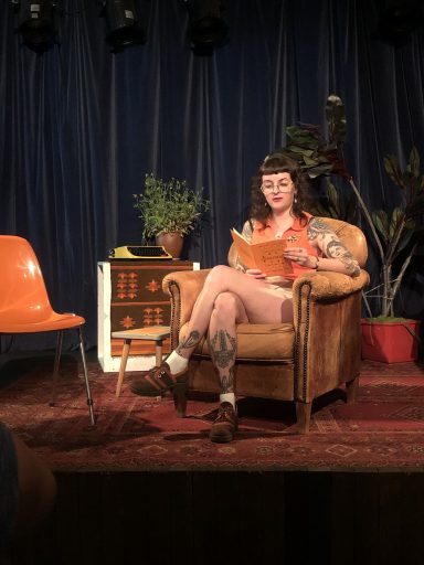 Vrouw met tattoos zit in een stoel, leest een boek in een sfeervolle ruimte.