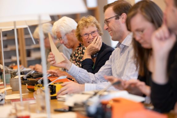Een groep mensen aan een lange tafel werkt met typemachines en papieren.