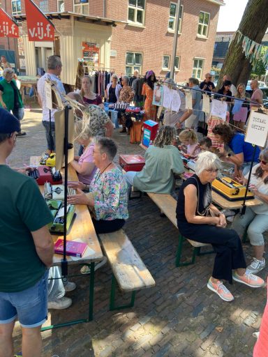 Drukke markt met bezoekers, tafels met standen en mensen die met elkaar converseren.