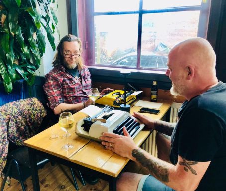Twee mannen aan een tafel met een typemachine en glazen wijn, binnen in een café.