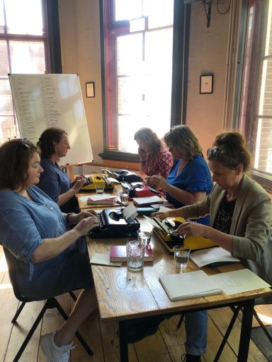 Vrouwen aan een tafel met typemachines, bezig met schrijven en aantekeningen maken.