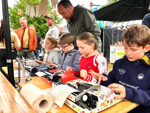 Kinderen typen op typermachines aan een tafel, terwijl volwassenen op de achtergrond kijken.