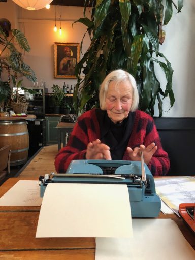 Oude vrouw met grijs haar typt op een blauwe typewriter, omgeven door planten.