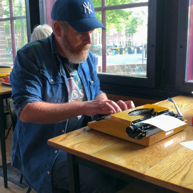 Man met baard typt op een gele typemachine aan een houten tafel in een café.
