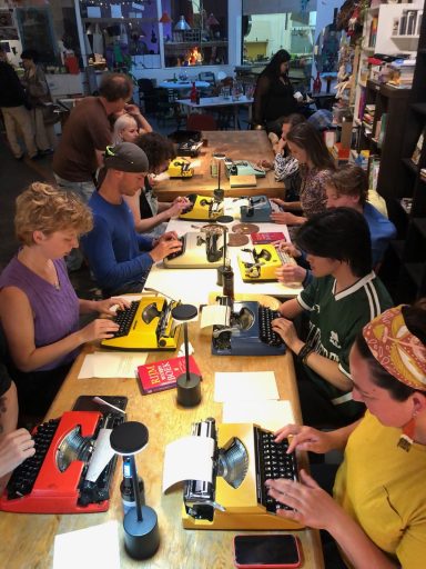Een groep mensen typt op oude typemachines aan een lange tafel.