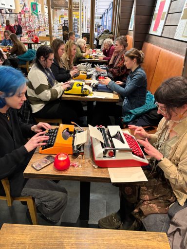 Groep mensen zit aan tafels in een café, typewriter en papieren voor zich.
