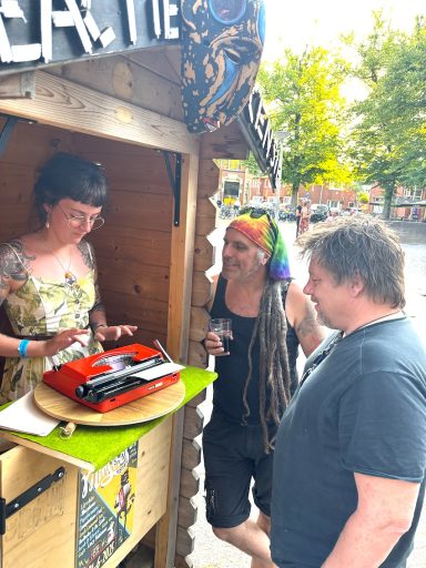 Een vrouw serveert taart aan twee mensen, één met dreadlocks en een regenbooghoed.