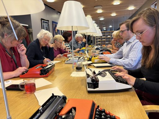 Een groep mensen typt op typemachines aan een lange tafel, met lampen boven hen.