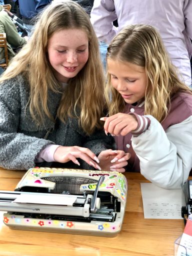 Twee meisjes glimlachen terwijl ze samen een typemachine gebruiken.