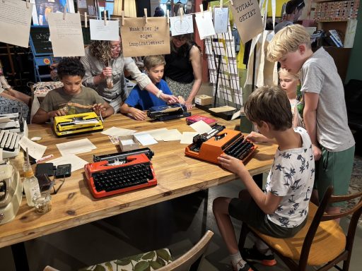 Kinderen experimenteren met typemachines aan een houten tafel in een interactieve ruimte.