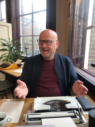 Man met een bril en een grijs jasje, zit aan een tafel met een typewriter en koffie.