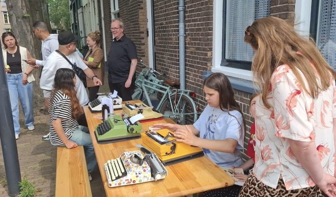 Groep mensen buiten aan een lange tafel met typemachines en fietsen op de achtergrond.